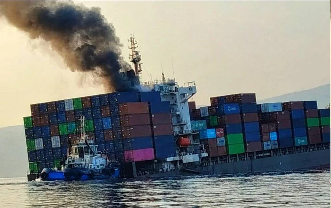 ontainer ship listing heavily in the water with thick black smoke pouring from its superstructure as a tugboat operates alongside.